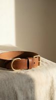 Brown leather dog collar with gold brass buckle and D-ring on linen surface, warm natural light, pet accessories concept.