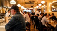 Busy restaurant interior with chef in kitchen overlooking dining room full of customers enjoying meals in warm atmospheric lighting.