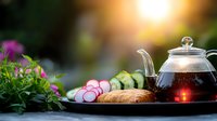 Healthy breakfast setting with glass teapot, fresh croissant, sliced radishes and cucumbers on dark tray against blurred garden background with morning sunlight.
