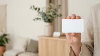 Person holding blank white card in modern home interior with potted plants and wooden furniture, suitable for business messaging and promotional content.
