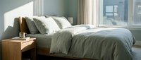 Cozy bedroom interior with wooden bed frame, soft green and white bedding, decorative pillows, wooden nightstand, and natural light from large windows with curtains.