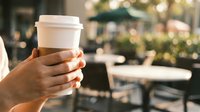 Person holding takeaway coffee cup outdoors in urban cafe setting with blurred background, suitable for beverage, lifestyle, and hospitality marketing materials.
