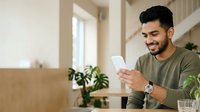 Young man with dark hair and olive complexion wearing casual olive-green shirt, smiling while holding smartphone in modern bright interior with potted plants and natural light from window.