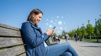 Young woman in denim jacket sitting on wooden bench using smartphone with social media icons floating above screen in sunny urban park setting.