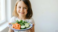 Happy young Caucasian girl holding plate of fresh vegetables including carrots and cucumbers, promoting healthy eating habits for children at home.