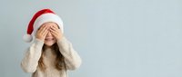Young woman in Santa hat covering eyes with hands in playful peek-a-boo gesture wearing cozy beige sweater against neutral background for holiday marketing.