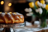 Sweet braided bread loaf dusted with powdered sugar on vintage metal cake stand, with soft bokeh lights and white tulips in background. For food blogs and bakery ads.