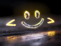 Glowing neon smiley face with bright yellow light effect floating above dark textured surface, creating warm atmospheric mood with bokeh reflections.