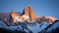 Majestic mountain peaks at sunrise with dramatic orange alpenglow illuminating snow-covered rocky spires against deep blue sky, creating stunning natural landscape.