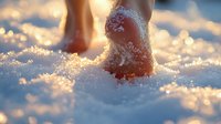 Bare feet walking in fresh powdery snow at sunset, creating footprints with golden sunlight illuminating crystalline snowflakes in a dreamy winter atmosphere.