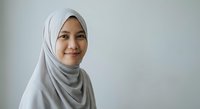 Young Muslim woman wearing gray hijab smiling warmly at camera against white background for professional headshot and business portrait use.