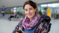 Young Caucasian woman wearing purple scarf and floral winter jacket smiling at airport terminal, casual travel style for cold weather destinations.