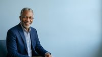 Senior African American male professional wearing navy blazer and striped shirt, smiling confidently against light blue background. Corporate portrait suitable for business websites, leadership profiles, and professional communications.