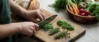 Woman chopping fresh herbs on wooden cutting board in kitchen with organic vegetables and bread for healthy cooking preparation.