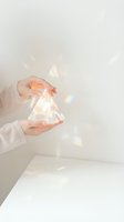 Hands holding transparent crystal prism with warm light refraction and rainbow bokeh effects on white background. Luxury cosmetics, fragrance, skincare packaging concept.