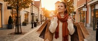 Young blonde woman enjoying autumn shopping spree with paper bags on charming cobblestone street during golden hour sunset in cozy sweater and scarf.