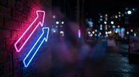 Bright neon arrows pointing upward on brick wall with blurred city lights background creating modern urban atmosphere for business growth concepts.