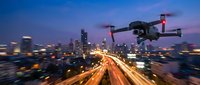 Modern drone flying over illuminated city highway at dusk with blurred traffic lights creating dynamic streaks against purple twilight sky and urban skyline.