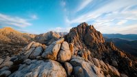 Dramatic rocky mountain peak at sunset with golden light illuminating granite boulders against vibrant blue sky with wispy clouds stretching across rugged wilderness landscape.