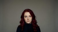 Professional portrait of confident young Caucasian woman with curly red hair wearing black turtleneck against neutral background for business headshots.