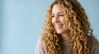 Happy young Caucasian woman with curly blonde hair smiling warmly in cozy sweater against soft blue background for lifestyle and wellness content.