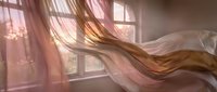 Woman with long brown hair flowing in wind near window with pink sheer curtains and warm sunlight, suitable for beauty, lifestyle and wellness design projects.