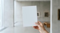 Person with medium skin tone holding blank white paper sheet in modern minimalist interior space with natural light for mockup and branding purposes.