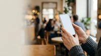 Person holding smartphone with blank white screen in modern office environment with blurred colleagues and holiday decorations in background.