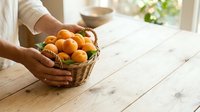 Hands holding wicker basket filled with fresh oranges on wooden table near window, ideal for food and harvest themes.