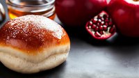 Fresh baked sweet bun with glossy brown top on dark background with red pomegranate and apples, selective focus on pastry with blurred fruits.