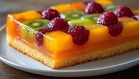 Colorful fruit tart with jelly layer featuring fresh raspberries, kiwi slices, and mango cubes arranged in geometric pattern on shortbread base, served on white plate.