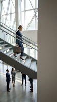 Professional businesswoman walking up modern office staircase while colleagues discuss below in contemporary corporate building with large windows.
