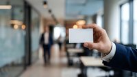 Businessman in dark suit holding blank white business card in modern office lobby with blurred colleagues and glass partitions in background for corporate branding and networking purposes.