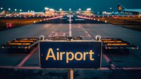 Airport runway at dusk with illuminated directional sign in foreground and landing lights creating leading lines into distance, moody aviation scene.