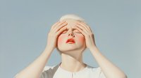 Young woman with pale skin and short blonde hair covering eyes with hands, wearing white top against light blue background, expressing emotional state.