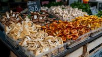 Fresh assorted edible mushrooms displayed at farmers market stall, including white button, shiitake and golden chanterelles arranged in wooden crates.