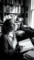 Middle-aged Caucasian woman writer contemplating in home office with vintage typewriter, bookshelves and natural window light in black and white.