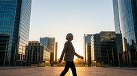 Businesswoman walking through urban downtown district at golden hour sunrise, surrounded by modern skyscrapers and glass buildings, representing career growth and professional success.