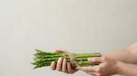 Fresh green asparagus bunch held in hands against neutral background, suitable for food, cooking, healthy eating, nutrition, and organic produce marketing materials.