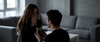 Young Caucasian man proposing marriage to surprised woman with engagement ring in modern living room. Romantic proposal moment at home.