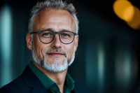 Confident mature Caucasian businessman with gray beard and glasses smiling at camera. Professional corporate portrait with blurred background and warm lighting.