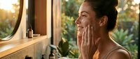 Young woman with medium skin tone applying skincare product to her face in a bright bathroom with natural window light and green plants visible outside, suitable for beauty, wellness and cosmetics marketing.