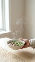 Person holding warm noodle soup bowl with steam rising, seated at wooden table near window with natural light for food and lifestyle content.