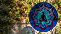 Stained glass mandala with meditation silhouette in lotus flower design featuring vibrant blues and teals against natural outdoor garden background.