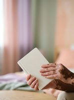 Close-up of hands holding blank white notebook against soft pastel striped background. Adult with medium skin tone displaying open journal for writing, planning, or note-taking purposes.