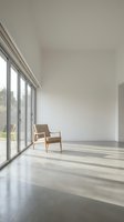 Minimalist modern interior with wooden chair by large windows, natural light casting shadows on polished concrete floor in contemporary living space.