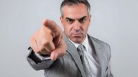 Serious middle-aged businessman in gray suit pointing accusingly at camera with stern expression against neutral background.