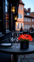 Romantic outdoor restaurant table setting with wine glasses and vibrant red geraniums in black pot against blurred European cityscape background at dusk.