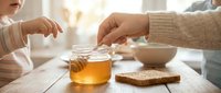 Two people sharing honey from a jar at a wooden table with toast and bowl in soft natural light.