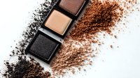 Crushed eyeshadow palette with scattered powder in brown, gold and black shades on white background, showing makeup texture and pigmentation for cosmetic advertising.